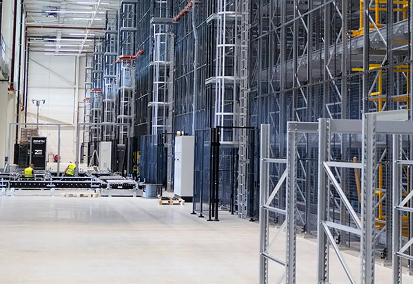 Spacious warehouse with tall metal racks, equipment, and workers in yellow vests