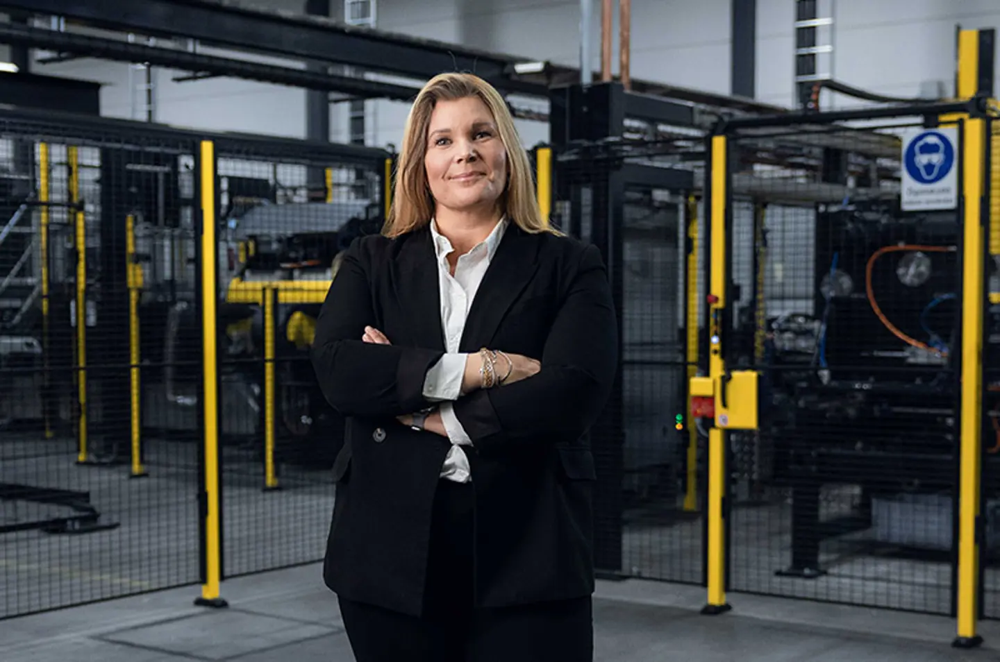 Die CEO von Axelent, Karin Sandén Ahlqvist, steht mit gekreuzten Armen vor einer Industriekulisse mit schwarzen Gitterwänden und gelben Pfosten